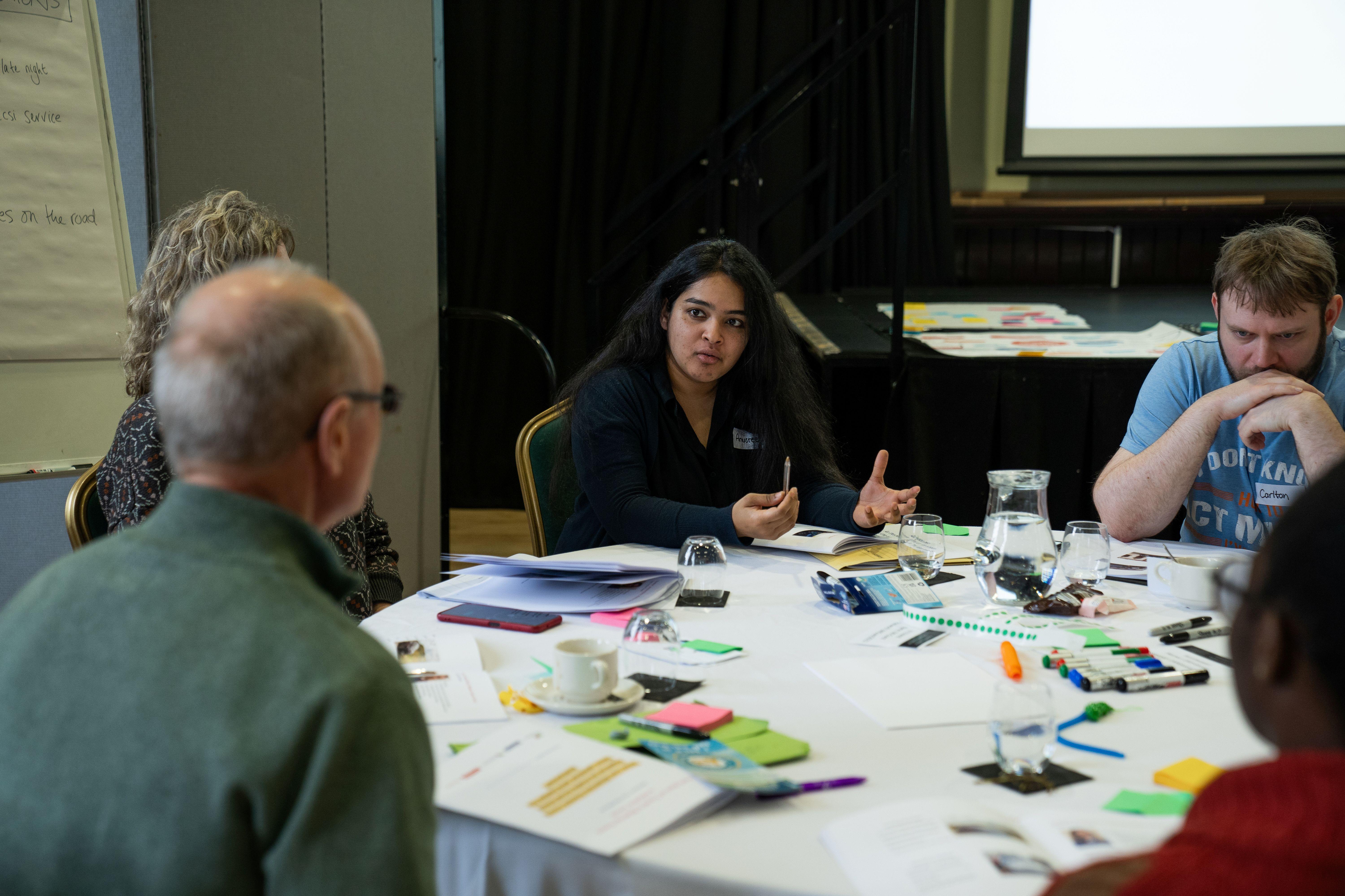 individuals sit around table discussing topic, the table is covered with pens, post-it notes and papers