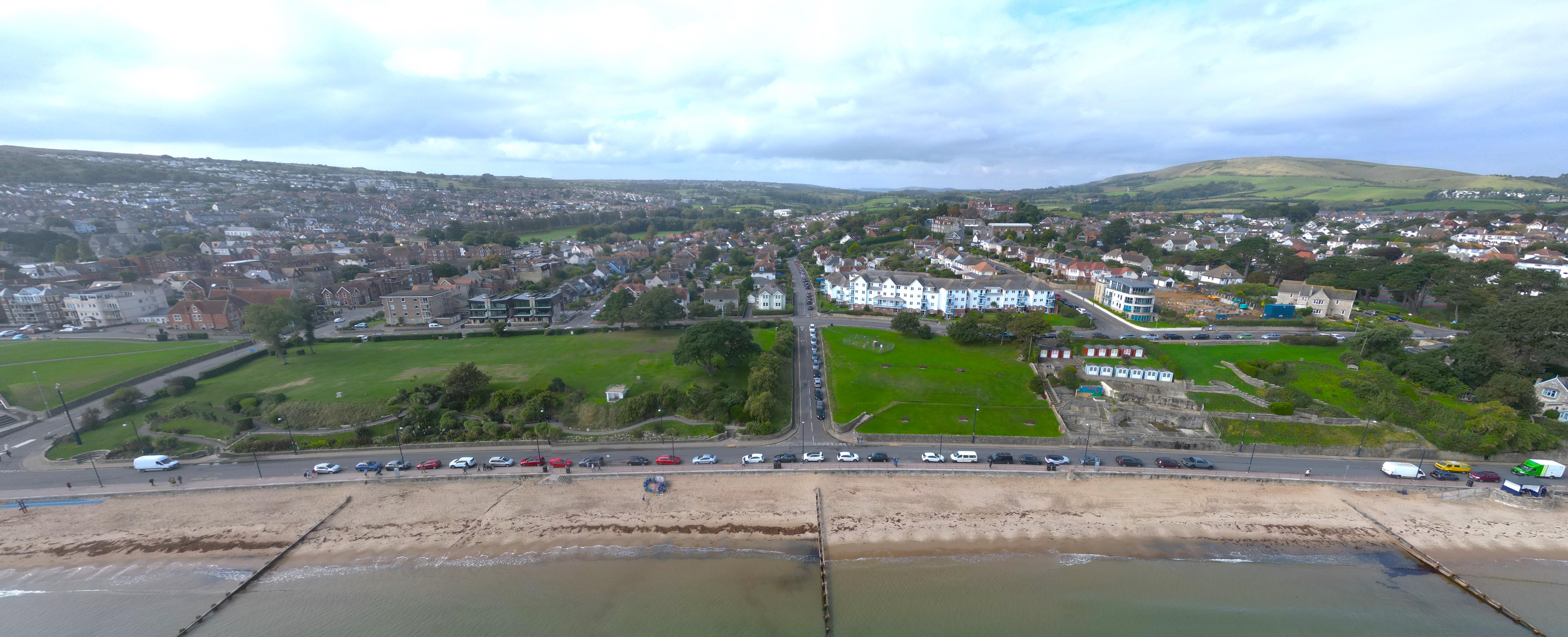 A road running parallel with a beach with green behind and further back there are buildings and hills and blue sky into the distance