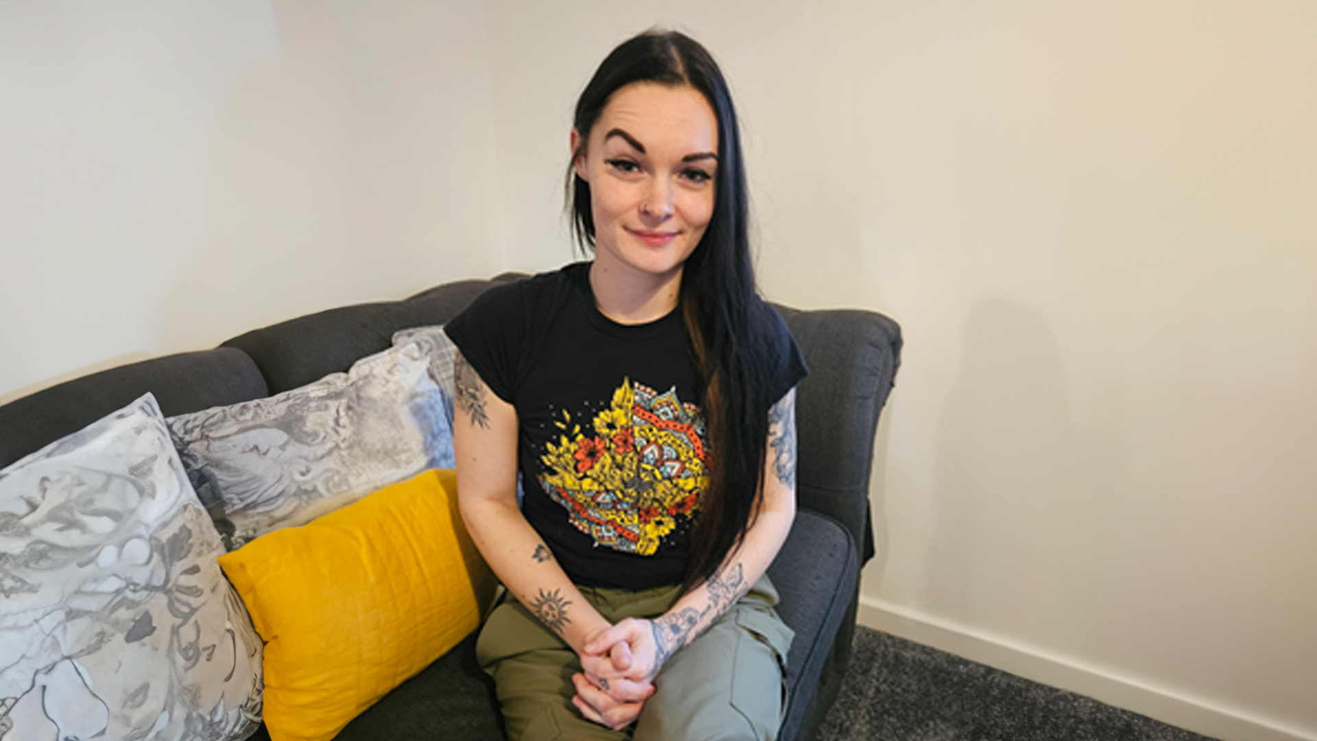 A white woman in her 20s and long black hair is sat on her grey sofa with cushions facing the camera.