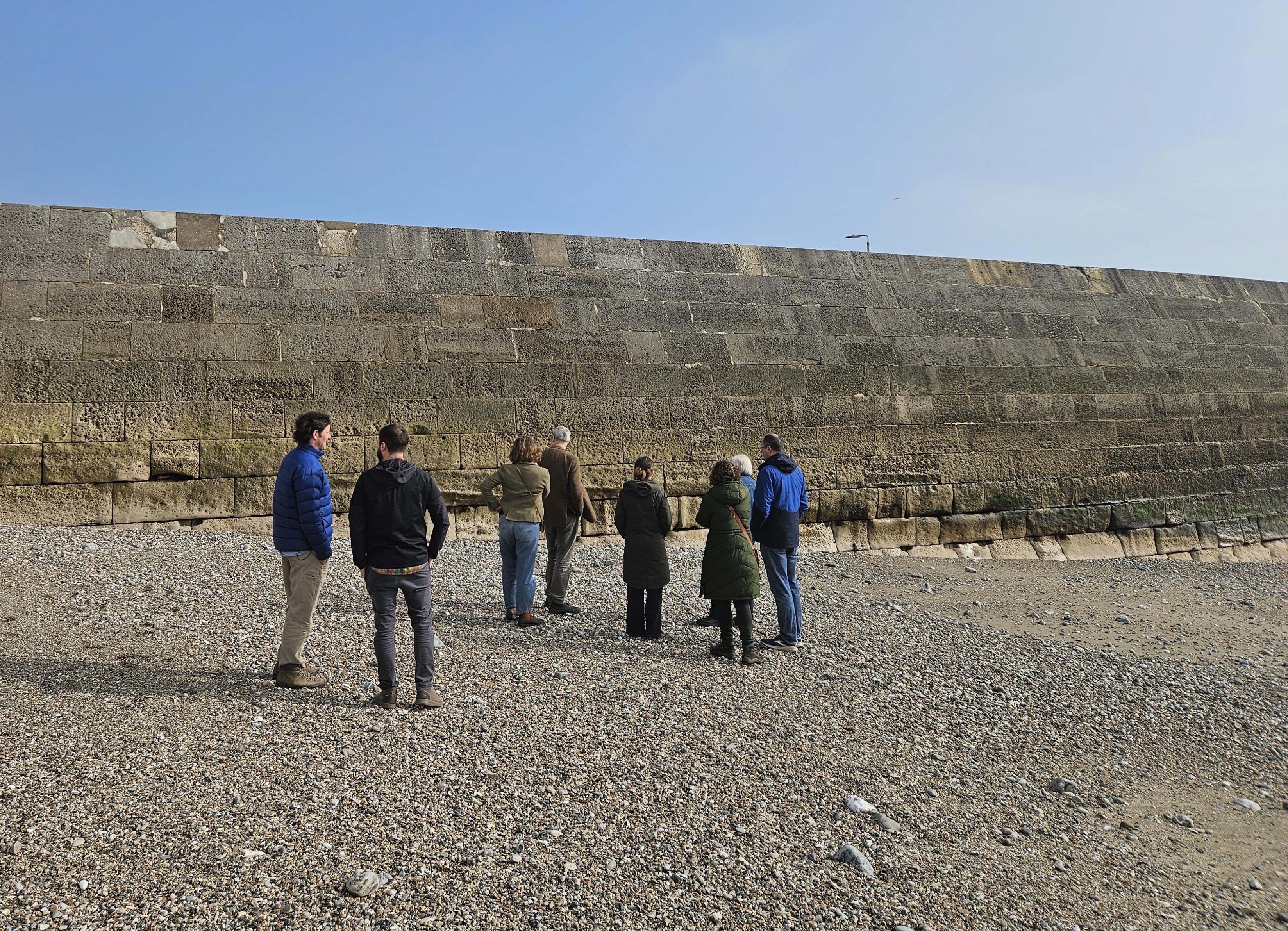 A group of people on a shingle beach looking at a wall structure