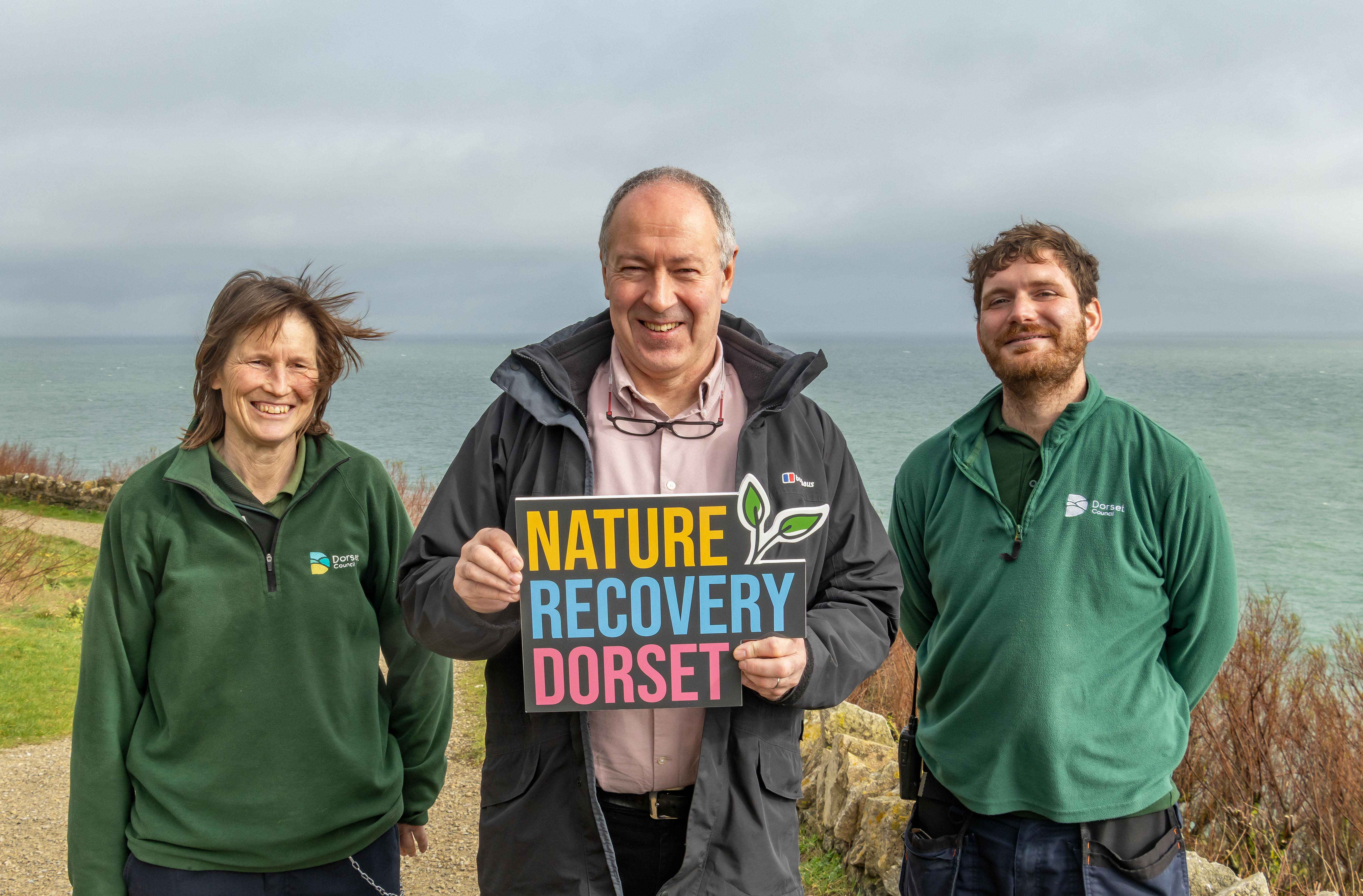 A woman wearing a green jumper standing next to a man in navy coat and another man wearing the same green jumper. The man in the middle is holding up a sign that says nature recovery Dorset. They are on a cliff top with the sea in the background