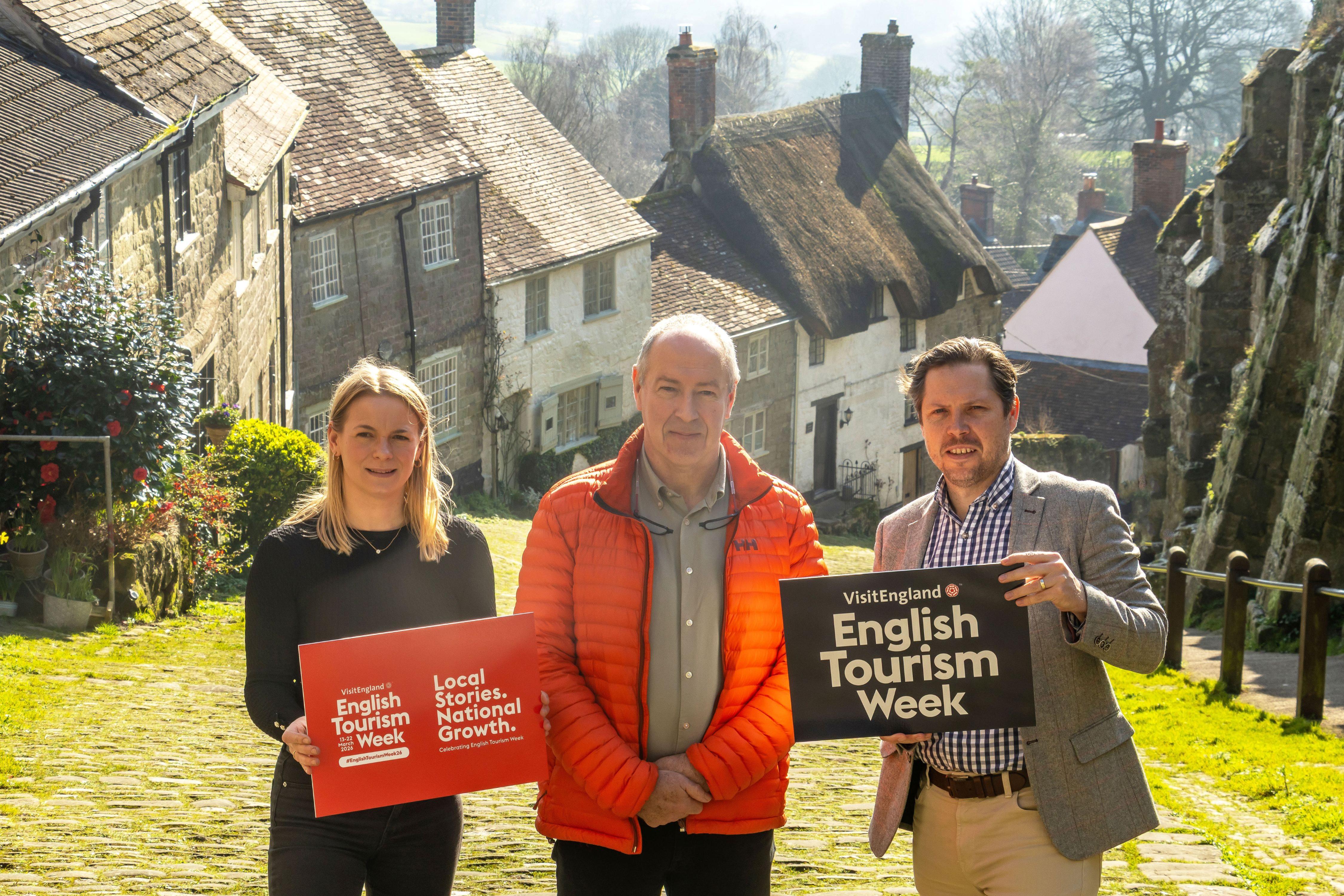 A woman with blonde hair holding a visit england sign, a man in middle wearing bright orange jacket in centre, man with tweed jacket holding visit england sign. All three standing on a cobbled path with thatched cottages in background.
