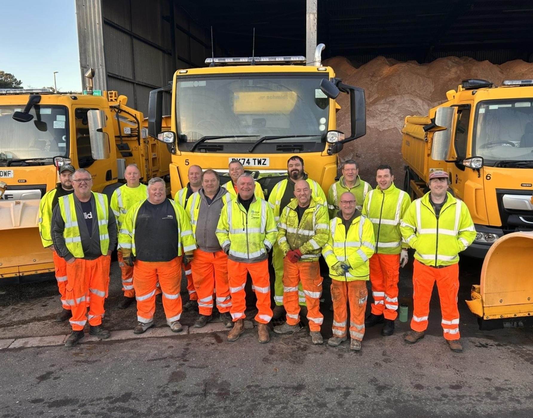 A line of up of some of the council's highways crew in front of some yellow gritting vehicles. They are wearing yellow jackets and orange trousers