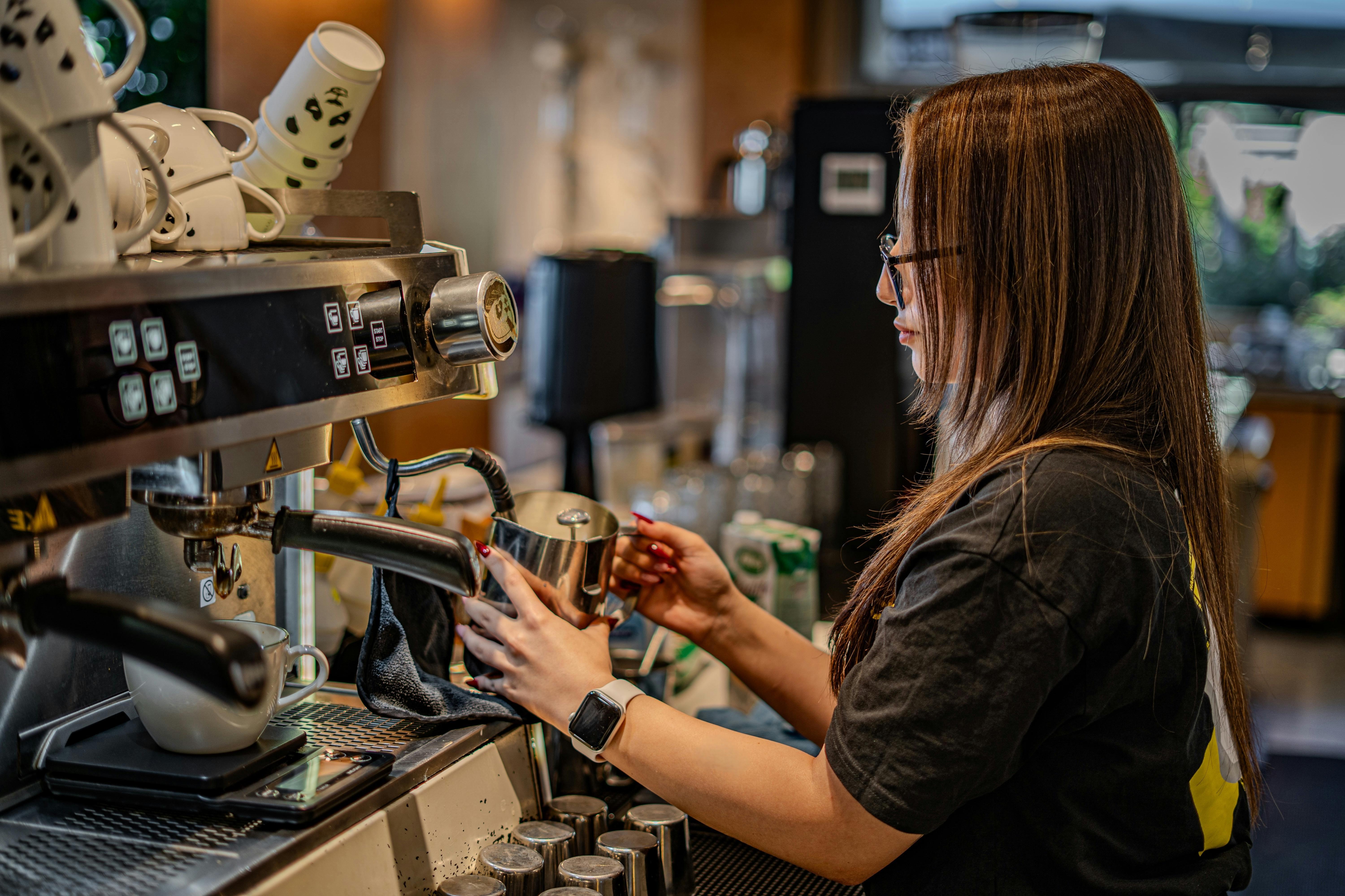 A young woman warming a jug of milk at a coffee machine