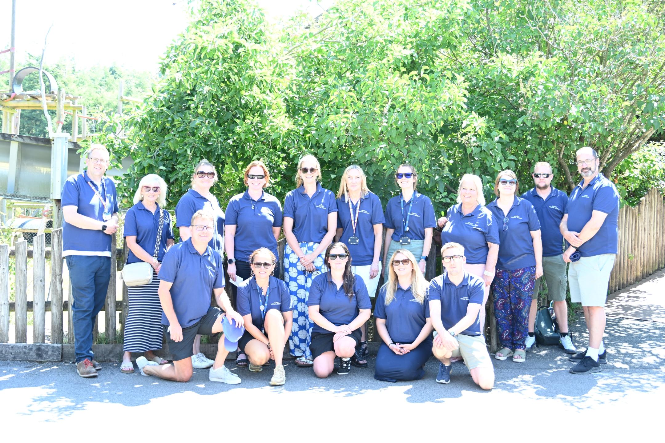 A group of around 16 adults wearing matching navy-blue polo shirts pose for a group photo outdoors on a sunny day. Some people are standing while others are kneeling in front. The group is gathered on a paved path in front of a wooden fence.