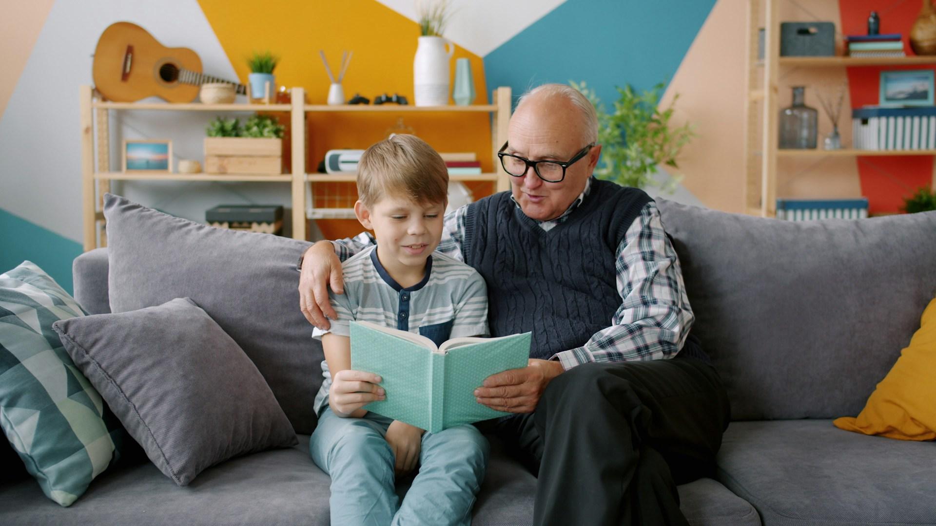 Older man reading to a child, sat on a couch with a book