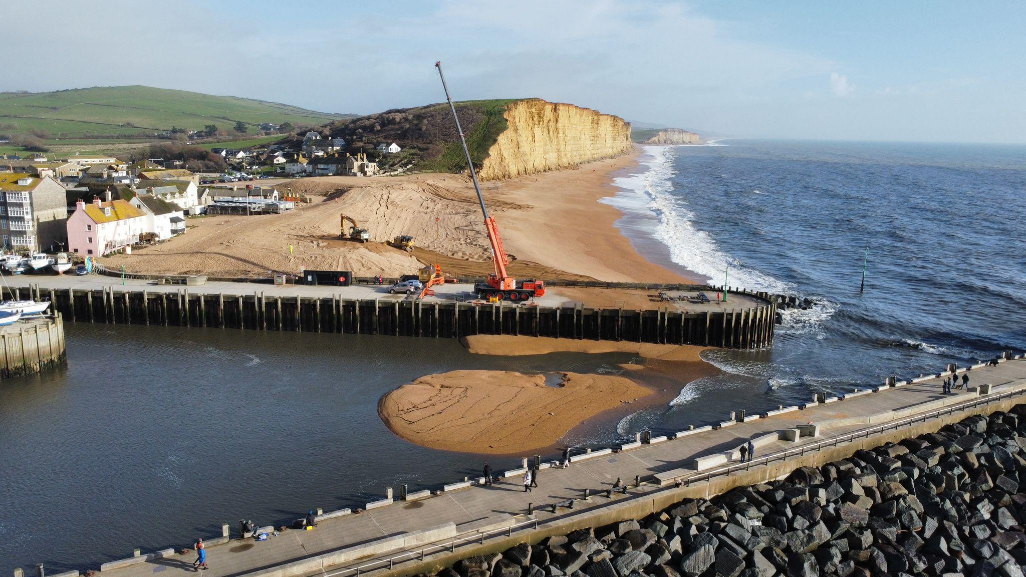 A drone shot of West Bay harbour, beach and cliffs. It shows the crane currently in place helping to clear away storm debris