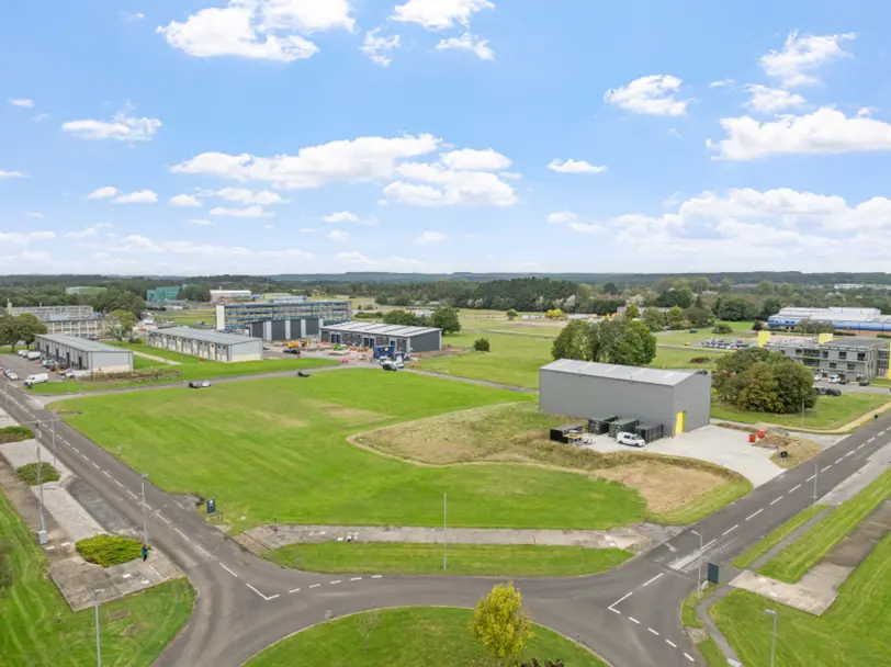Aerial view of a business or industrial park with several large buildings, open green spaces, access roads, and surrounding woodland under a bright sky.
