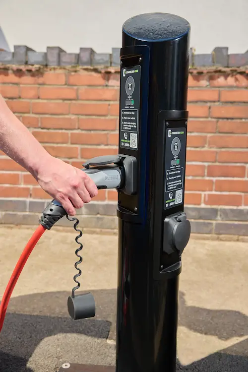 Hand plugging a charging cable into a black Connected Kerb electric vehicle charging unit beside a brick wall.