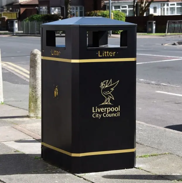 Black metal litter bin housing with gold bands and lettering, square shape with covered top and side openings, placed on a paved high street.