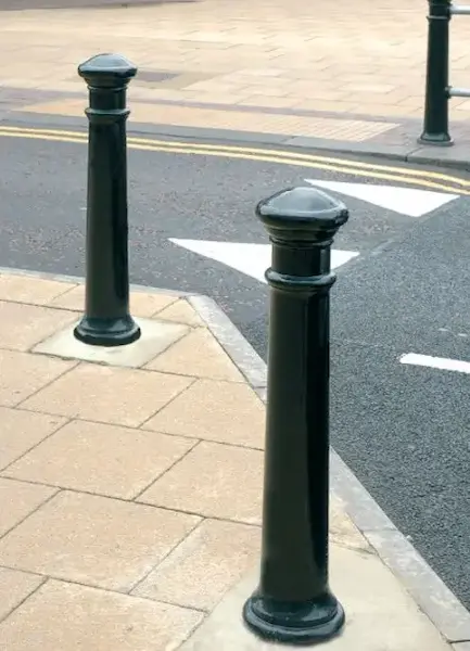 Black cast iron bollard with a rounded domed top and decorative detailing, installed along a pavement to separate pedestrians from the road.