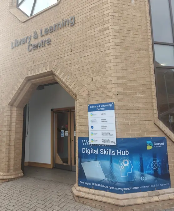 Entrance to a brick Library and Learning Centre with signage for services and a banner promoting the Weymouth Digital Skills Hub.