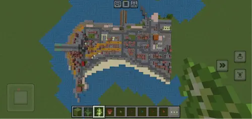 Top‑down view of a Minecraft build showing a detailed coastal town layout with roads, buildings, and waterfront areas surrounded by water and green land.