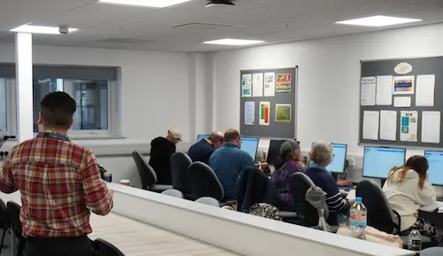 Classroom scene with several people seated at computers while an instructor stands at the front of the room.