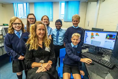 A classroom scene showing a group of students in school uniforms gathered around a teacher at a computer workstation, with coding software displayed on the monitor.