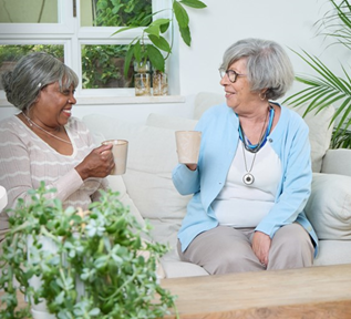 Two ladies sat on a sofa, smiling at each other whilst holding coffee cups.