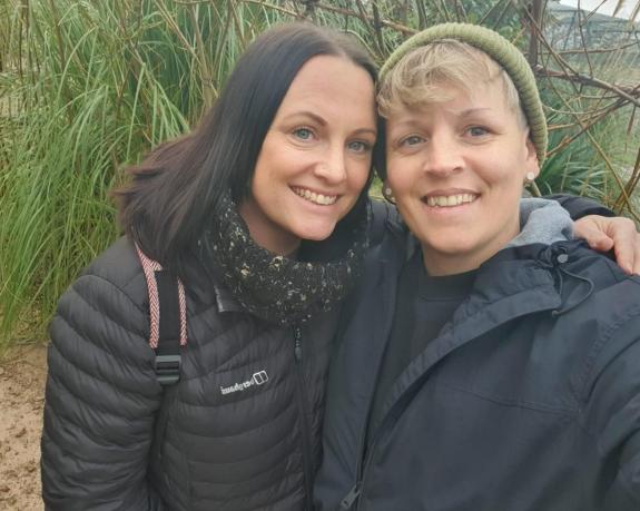 Keri and Loren smile at the camera happily. They are both wearing warm coats and appear to be standing on the beach.