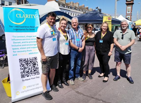 6 members of CLARITY, Dorset's new Lived Experience Recovery Organisation (LERO) in front of promotional stand on Weymouth seafront.