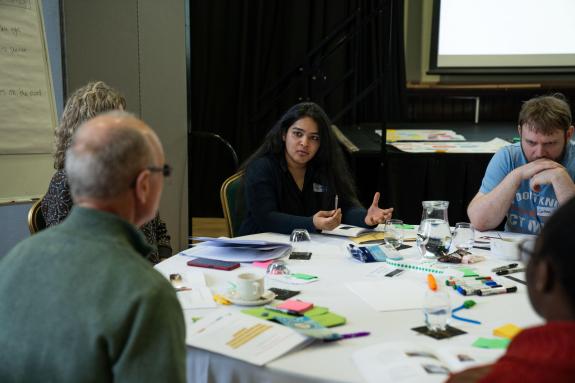 individuals sit around table discussing topic, the table is covered with pens, post-it notes and papers