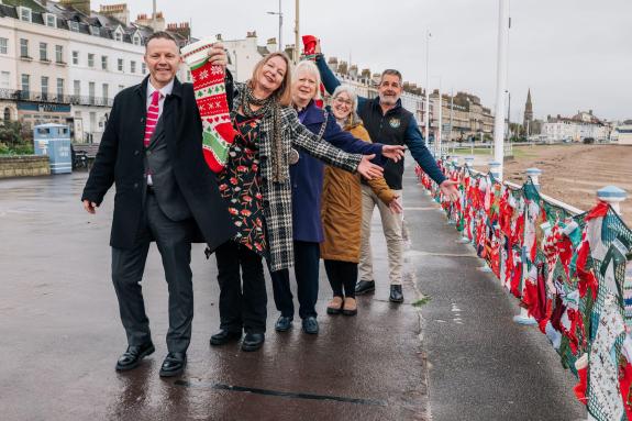 Five people stand proudly in front of a large display of Christmas stockings. They are on display along Weymouth Esplanade to raise awareness of fostering