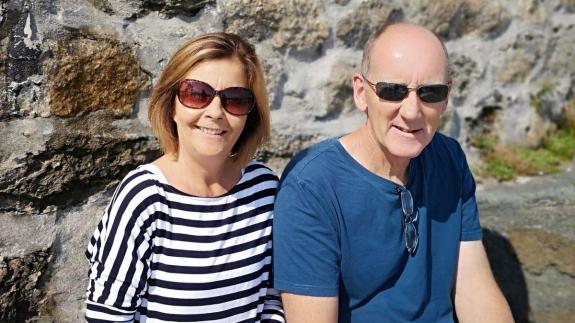 Sallyanne and Paul smile at the camera in the sunshine. They foster for Dorset Council. Sally has a stripe tshirt on and Paul is wearing a blue tshirt. Both appear very happy.