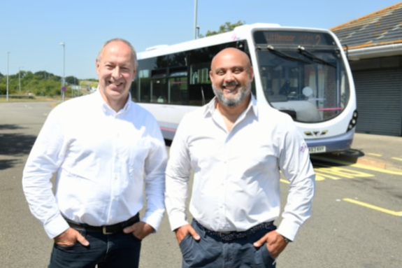 Two middle-aged men wearing white shirts and dark trousers stand with hands in pockets in front of a single decker First Bus on a Park and Ride site.